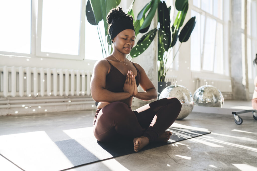 Young Black female athlete sitting cross-legged on a yoga mat in a spacious, light-filled studio, hands in prayer position and eyes closed, meditating in a moment of calm and focus.