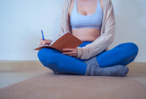 Young woman sitting cross-legged on a yoga mat, writing in a notebook during a quiet moment of self-reflection and planning.
