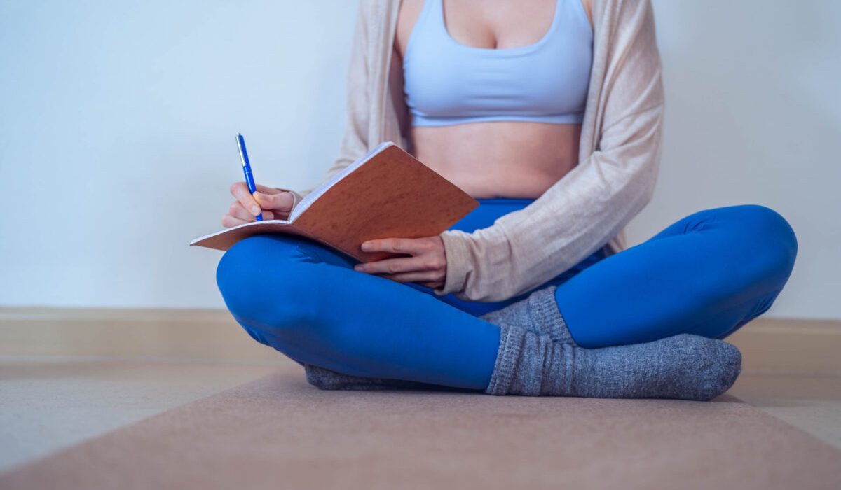 Young woman sitting cross-legged on a yoga mat, writing in a notebook during a quiet moment of self-reflection and planning.